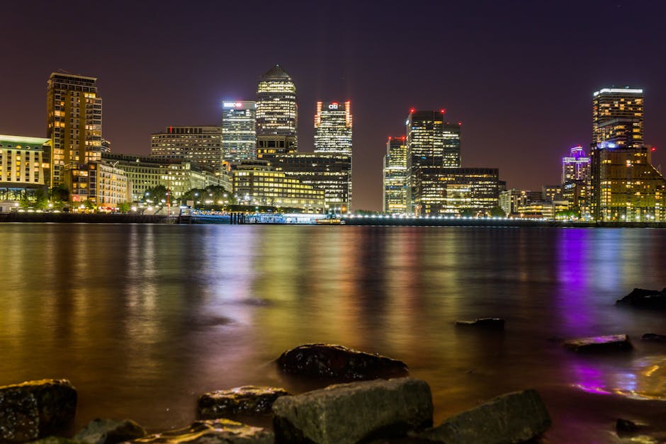 Nighttime cityscape of Canary Wharf viewed across the River Thames, with illuminated high-rise office buildings reflecting on the water. The buildings feature modern glass facades and are topped with colorful lighting, including red and purple accents. The scene is taken from the riverbank, where large rocks and stones are visible in the foreground, with the water appearing calm and slightly rippled due to long exposure. Interior lighting from buildings creates a vibrant glow, and the sky is dark, emphasizing the lit structures. This image relates to house and office removals, illustrating the logistics of furniture transport and packing processes in an urban environment, relevant to removals services provided by Removal Company Docklands for clients in the Docklands area, especially around Canary Wharf.