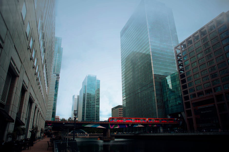 A street scene in Docklands featuring modern glass skyscrapers and office buildings. In the foreground, there are several cardboard boxes, some wrapped in plastic or fabric protective covers, placed on the pavement near an entrance. A trolley loaded with additional boxes and packing materials is positioned close to the buildings. A red train runs along an elevated track above the street, with part of the bridge structure decorated in colourful graphics. The image shows a typical urban environment suitable for house or office removals, captured during daylight hours with natural lighting. Removal Company Docklands may assist with furniture transport and packing during home relocations within this area, as indicated by the presence of moving equipment and packaging materials in the scene.