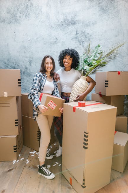 Two women are standing inside a room surrounded by numerous packed cardboard moving boxes, some with red packing tape, indicating a home relocation process. One woman, with curly dark hair, is holding a large decorative vase with green plant foliage, while the other woman, with straight brown hair, is carrying a smaller box decorated with red and white tape. The room has a textured grey wall and wooden flooring, with a mix of natural and artificial lighting. Several boxes are stacked to either side and behind them, suggesting careful packing for a furniture transport or moving service. The scene captures the final stages of packing or loading in a house removal, with visible packaging materials and the women’s expressions indicating they are preparing for the next step in the move. This visual context aligns with themes of house removals, packing, and moving logistics, as handled by Removal Company Docklands, serving the Isle of Dogs area for efficient home relocation services.