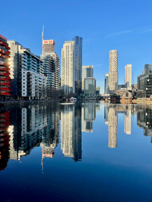 A wide view of a modern city skyline with tall office and residential buildings featuring glass facades, situated along the banks of a calm river that reflects the structures and the clear blue sky above. The buildings vary in height and design, including some with unique architectural features, and there are cranes visible on the rooftops indicating ongoing construction. The scene is taken during daylight with bright natural lighting, and the environment appears clean and orderly, highlighting the urban landscape of the Isle of Dogs area in London. This image exemplifies the type of scenery seen during home relocation or furniture transport projects in the Docklands, where removals specialist companies like Removal Company Docklands often operate.