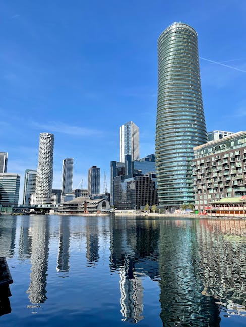 A clear daytime view of the Docklands cityscape featuring modern high-rise buildings, including a prominent cylindrical tower with glass windows reflecting the sky, and other tall structures with vary